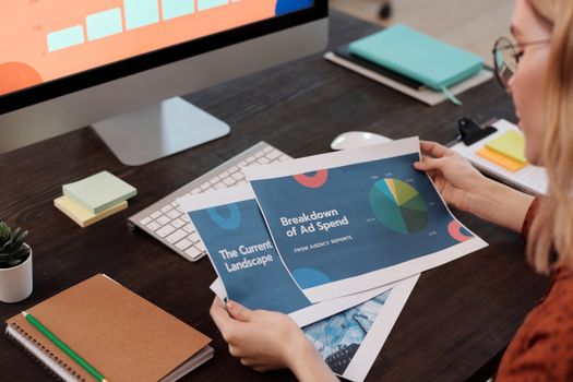 A businesswoman reviews marketing documents with charts at her desk, featuring a computer and office supplies.