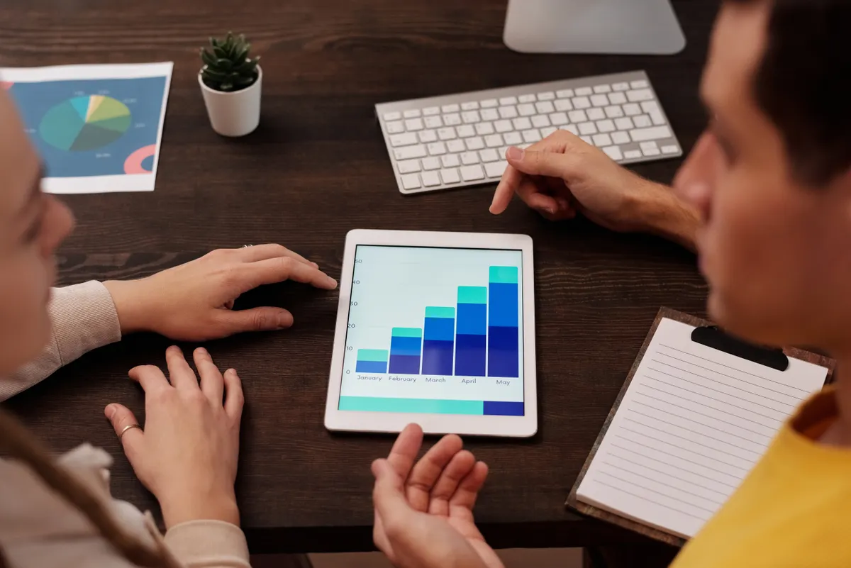 Two people at a desk reviewing a bar chart on a tablet, with a printed pie chart and a notepad beside them