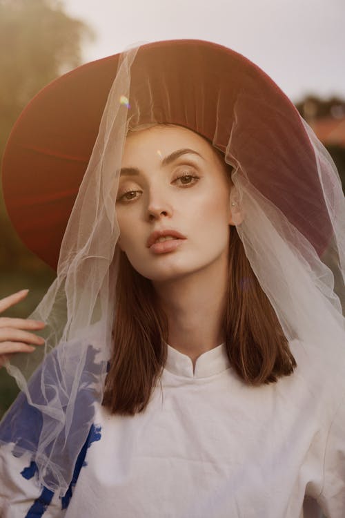 Charming female wearing broad brim with thin transparent veil in nature and looking at camera