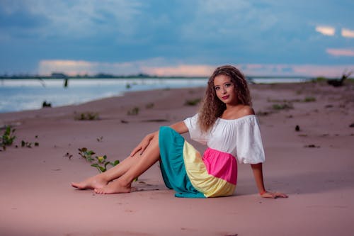 Woman in White Tank Top and Colorful Skirt Sitting on Brown Sand