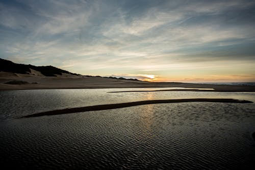 Sandy rough coast washed by tranquil rippling water of lake under cloudy sky in sunset