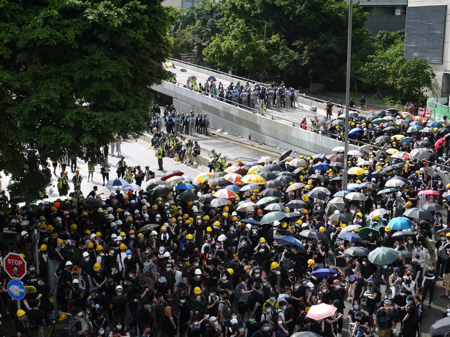 A high-angle view of a large protest with people holding umbrellas, facing police barricades on a city street.