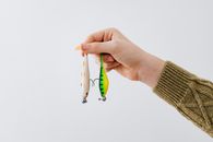 A close-up of a hand holding two colorful fishing lures against a white backdrop.