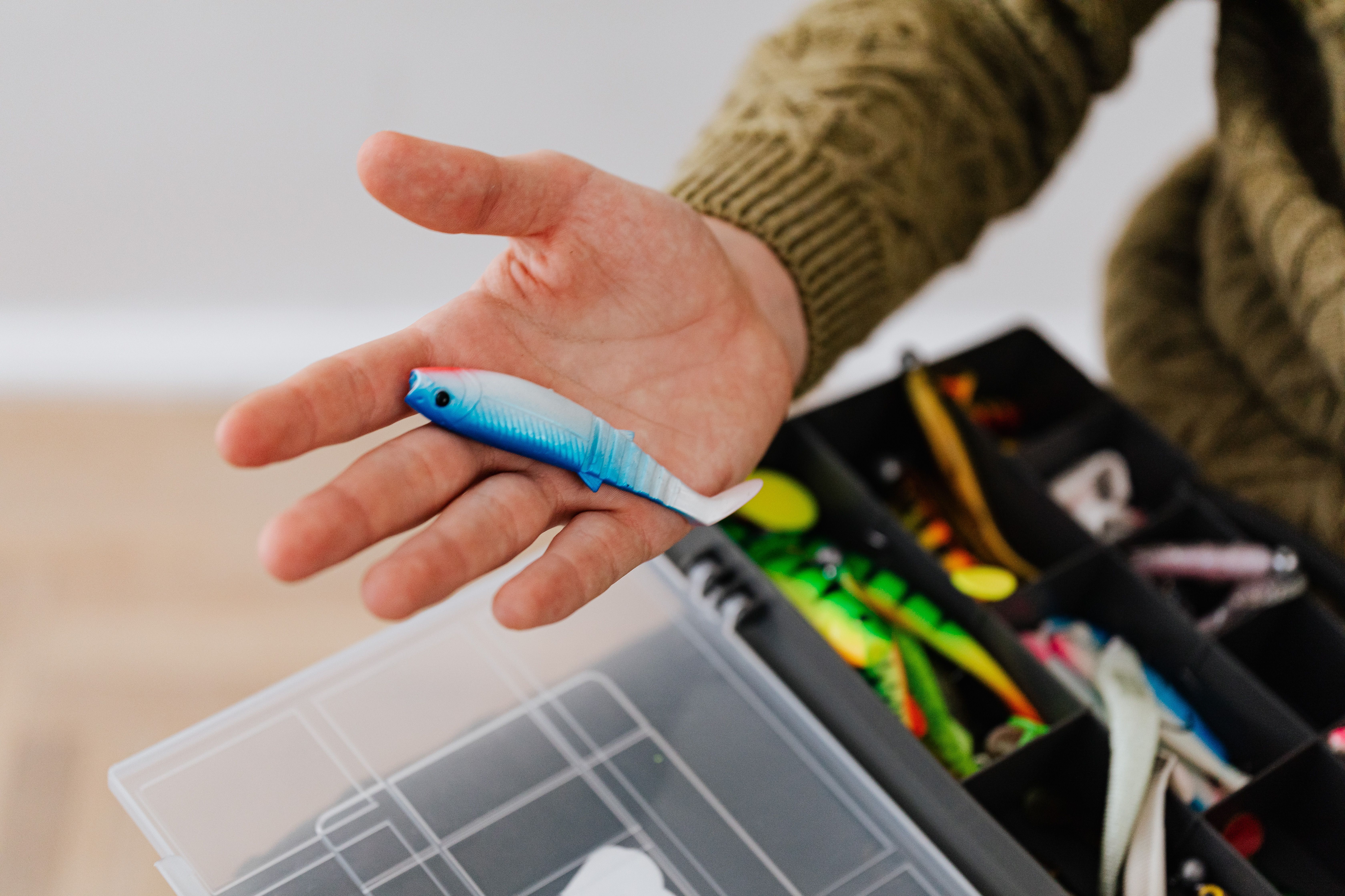 Close-up of a hand holding a fishing lure over a tackle box indoors.