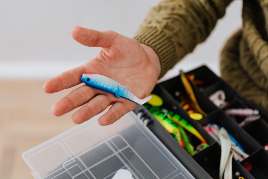 Close-up of a hand holding a fishing lure over a tackle box indoors.