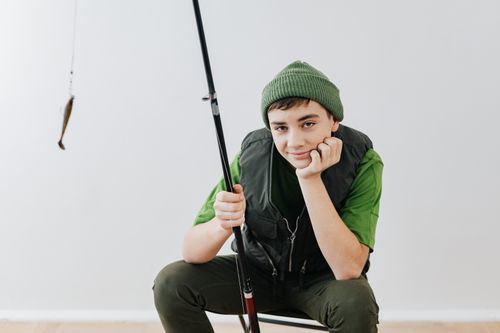 Free Smiling teenager in green beanie holding a fishing rod indoors, enjoying a leisure moment. Stock Photo