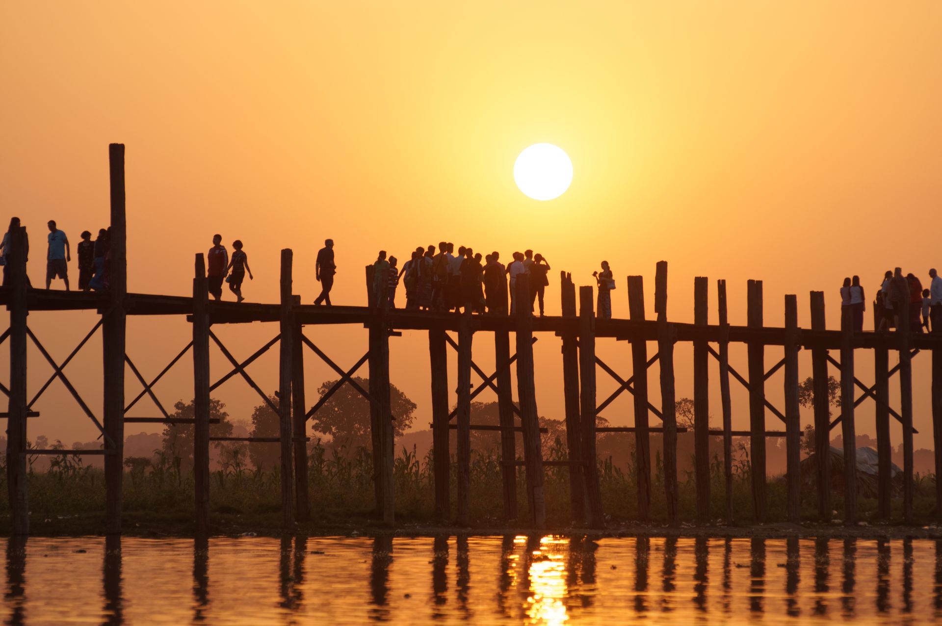 Backlit silhouettes of people crossing the U Bein Bridge during a golden sunset.
