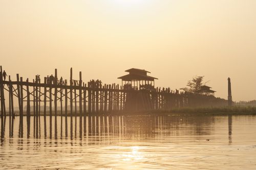 Free Silhouette of people walking on a wooden bridge at sunset with reflective water below. Stock Photo