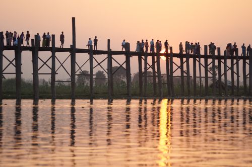 Free A serene sunset scene of people silhouetted on a wooden bridge with a reflection on the water. Stock Photo
