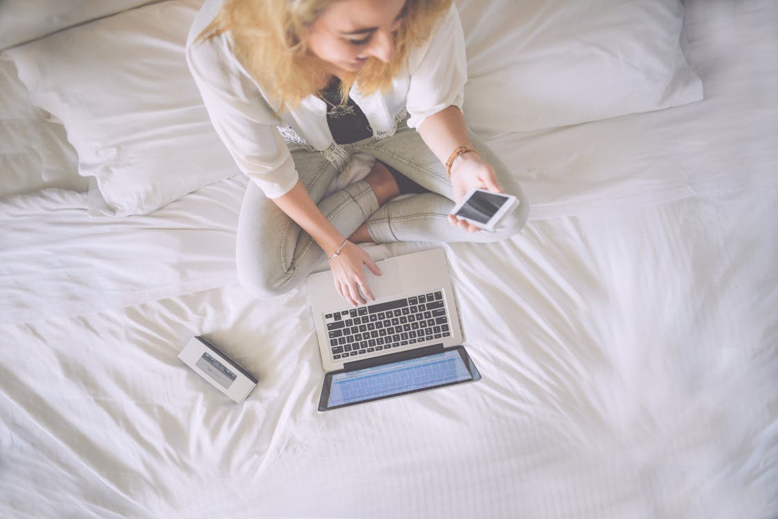 Free Woman Sitting on Bed While Holding Phone and Touching Macbook Pro Stock Photo