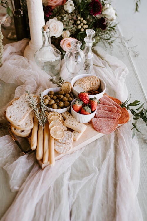 From above of wooden tray with assorted food for guests consisting of salami olives strawberries in bowls with bread and rosemary twigs on festive table