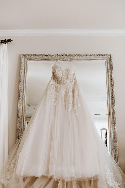 Low angle of elegant white bridal dress hanging on big framed mirror in light room before wedding ceremony