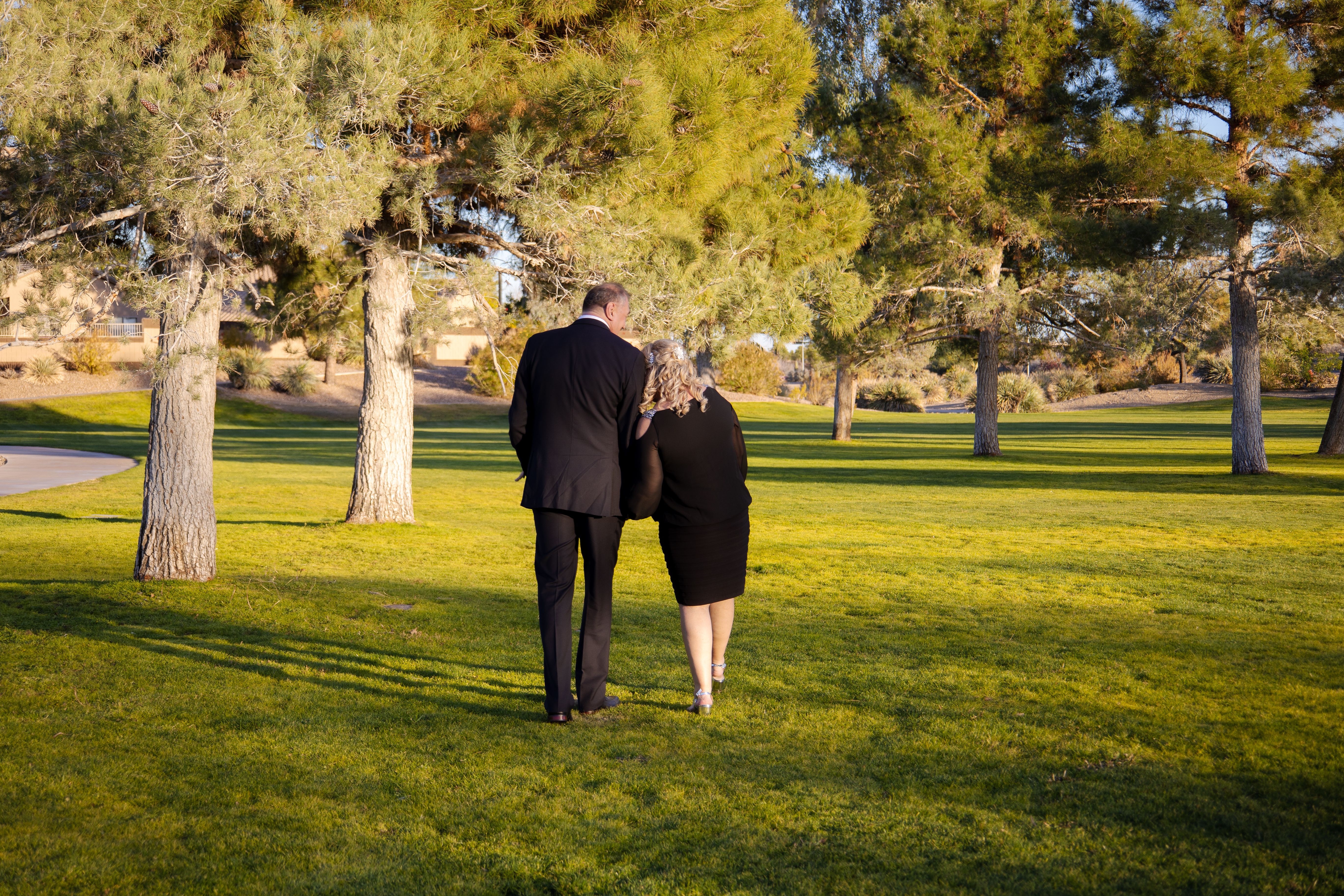 A couple walking arm-in-arm in a sunny park, surrounded by trees and grass.