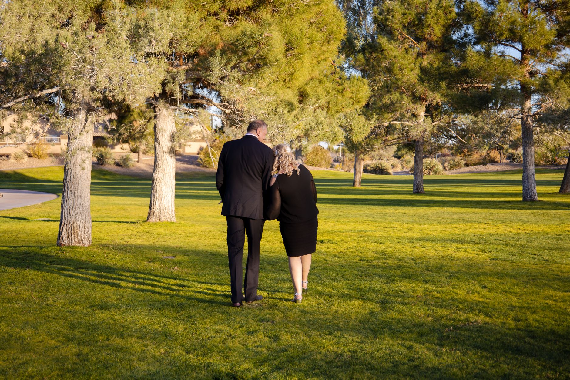 A couple walking arm-in-arm in a sunny park, surrounded by trees and grass.