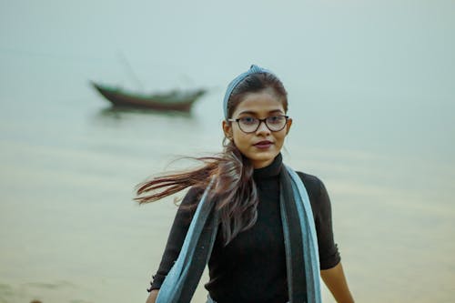 Calm Indian teenage girl in eyeglasses looking at camera while standing on coast near sea with shabby boat on blurred background