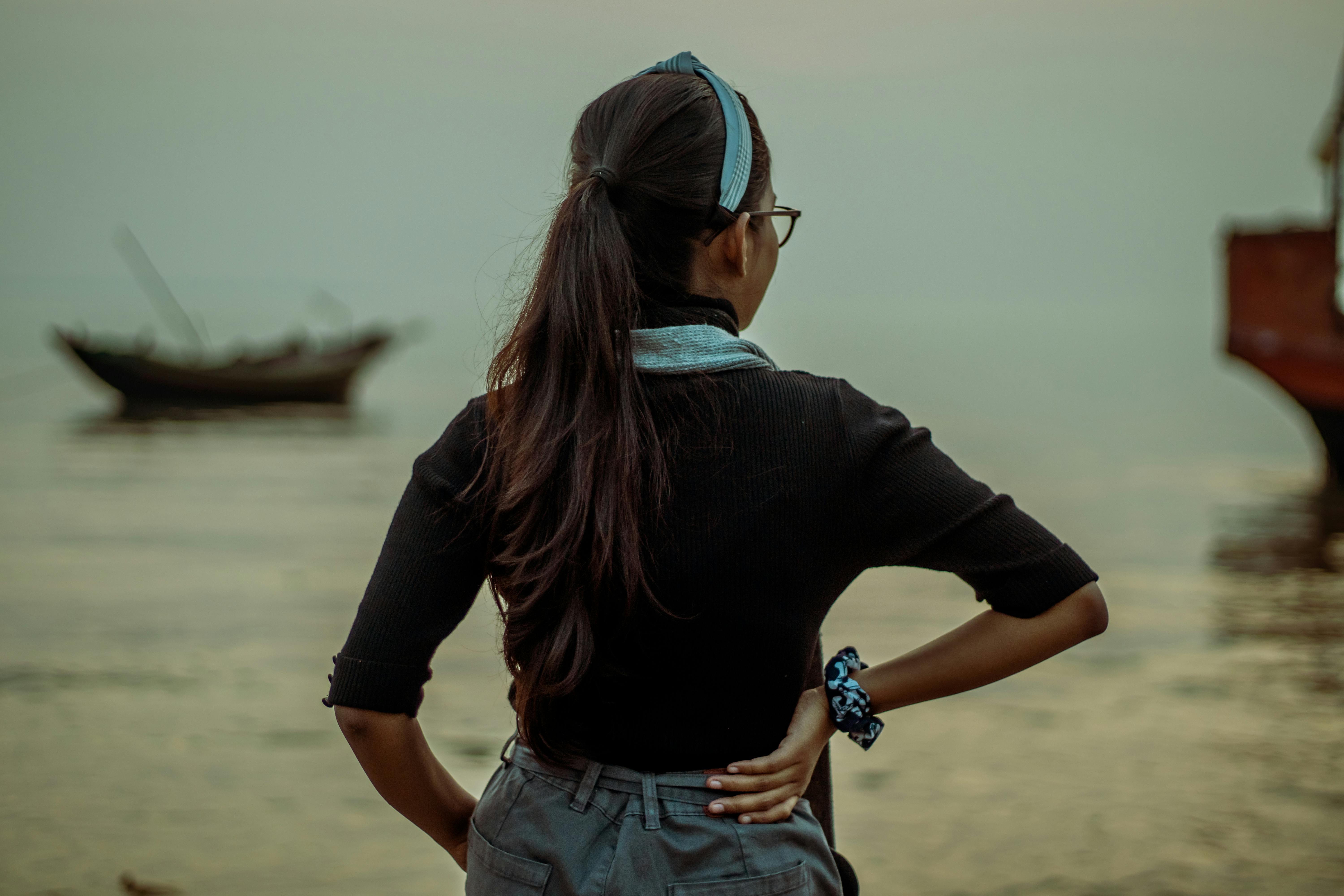 Back view of unrecognizable female with long dark hair standing on coast near sea with shabby vessels in coastal area