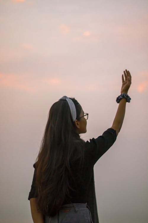 Side view of unrecognizable teenage girl with long hair standing against sundown sky with raised arm in nature on evening time