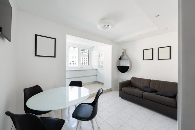 Interior of modern light house with glass table near chairs and couch with pillows near paintings and shelves on wall near entrance to kitchen with counter with window