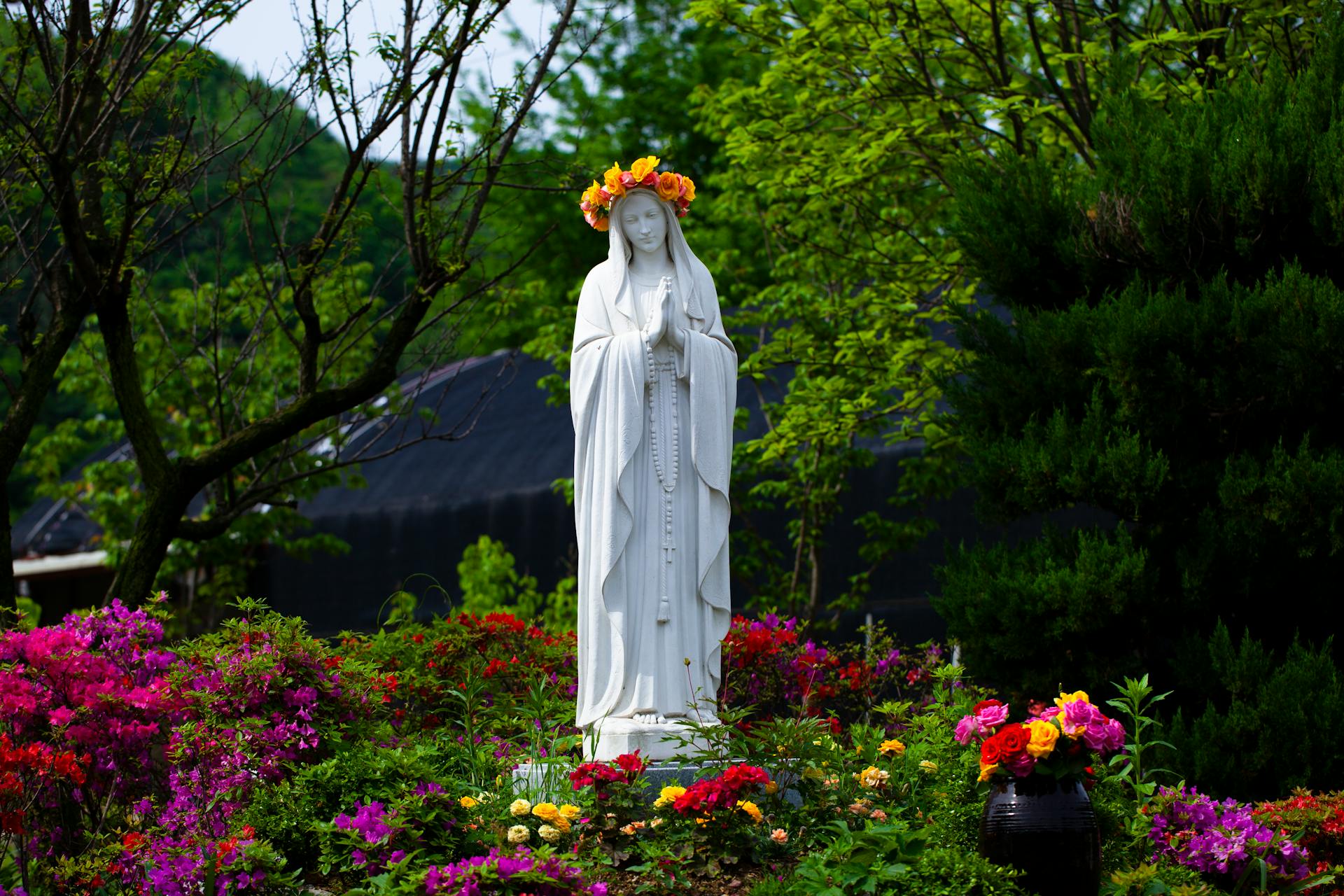 Statue of Virgin Mary adorned with a flower crown surrounded by vibrant garden blooms.