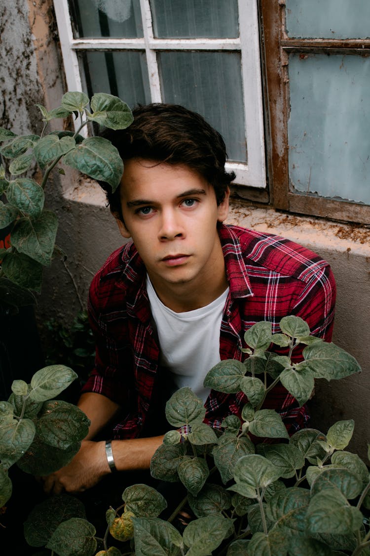 Serious Young Man Sitting Near Old House And Plants