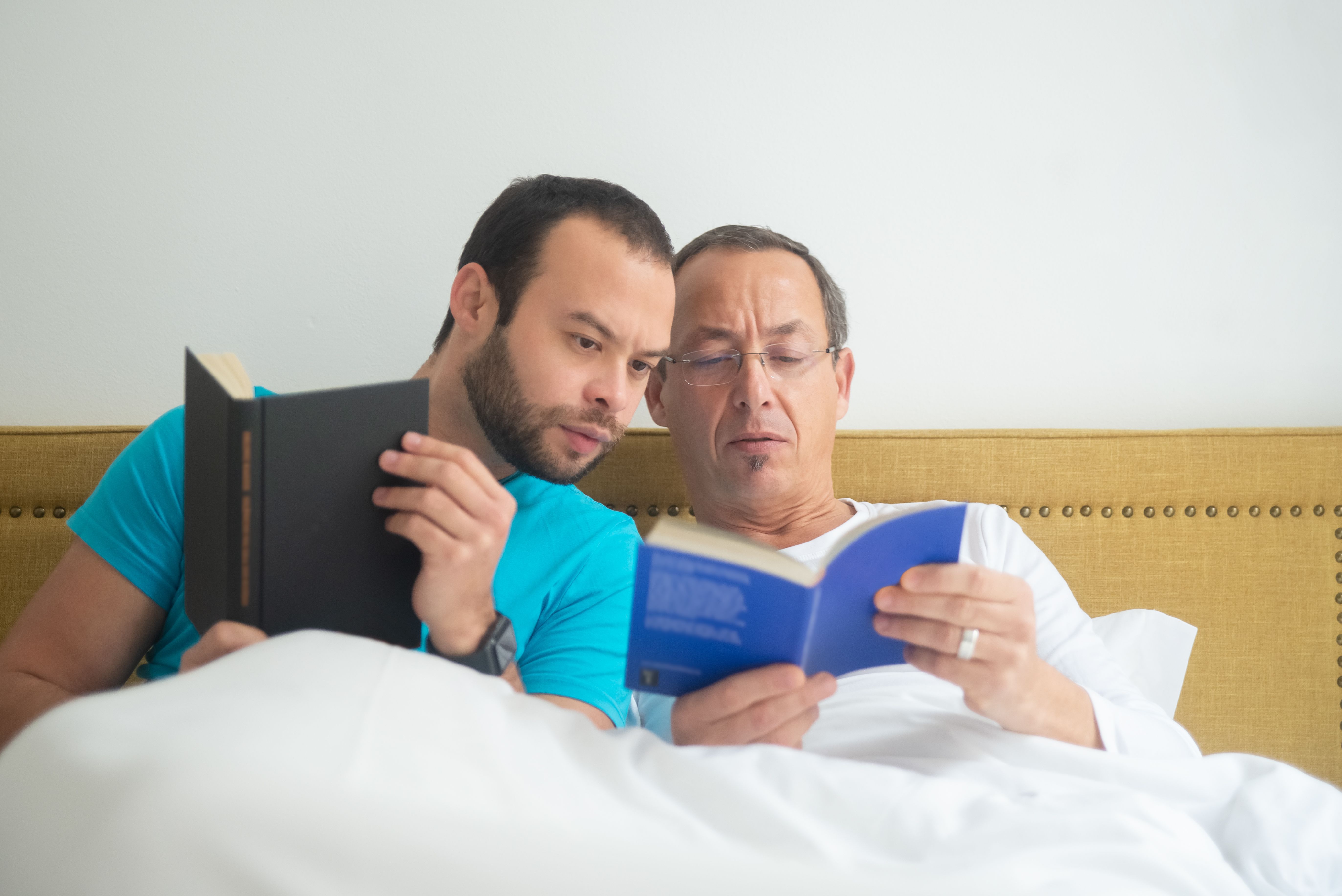Two men enjoying a peaceful reading moment in bed, representing love and relaxation.