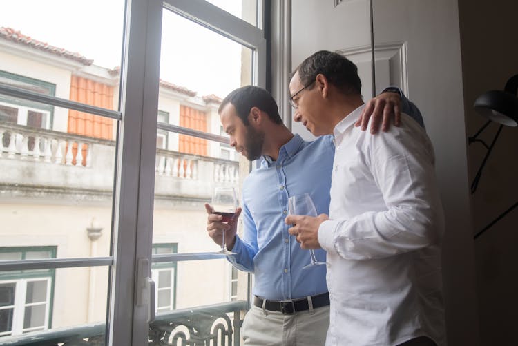 Two Men Standing By The Window Holding Wine Glasses And Looking Down