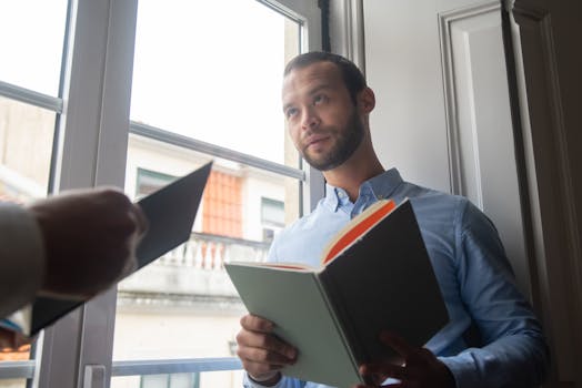 Man reading book by the window, thoughtful expression, indoor lighting.