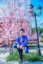 Portrait of a Boy Sitting on a Bench by a Street Lamp and a Pink Blossoming Tree