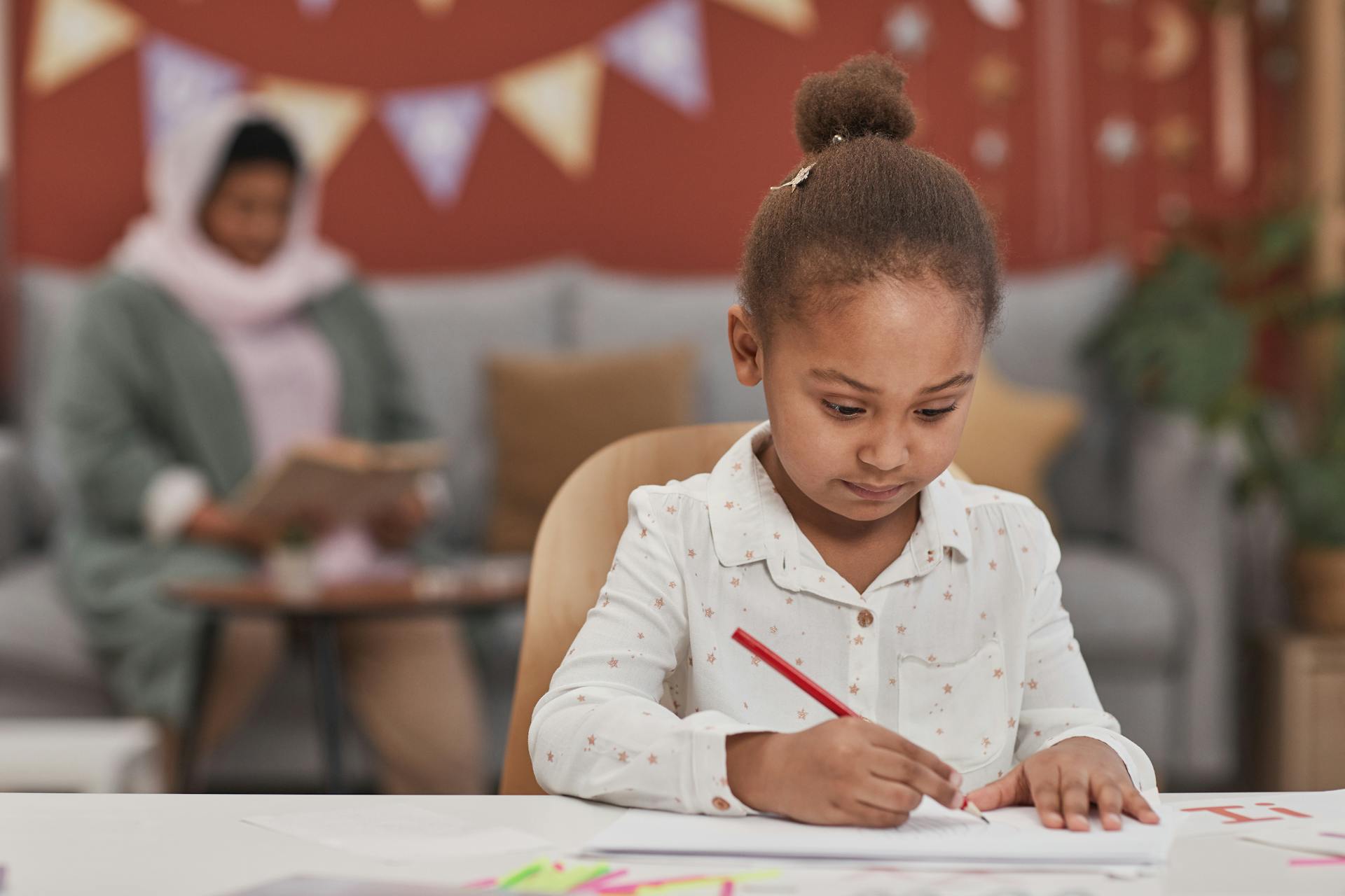 A young girl focuses on writing at a table, with an adult supervising in the background.