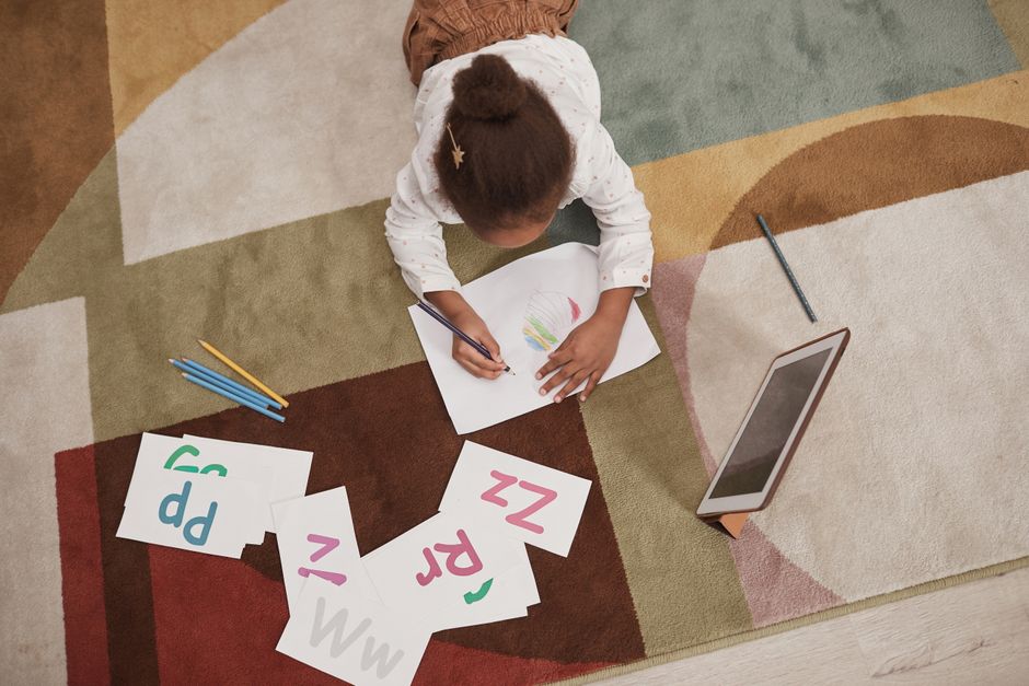 Young girl drawing letters with colored pencils on a patterned carpet, using a tablet for learning.