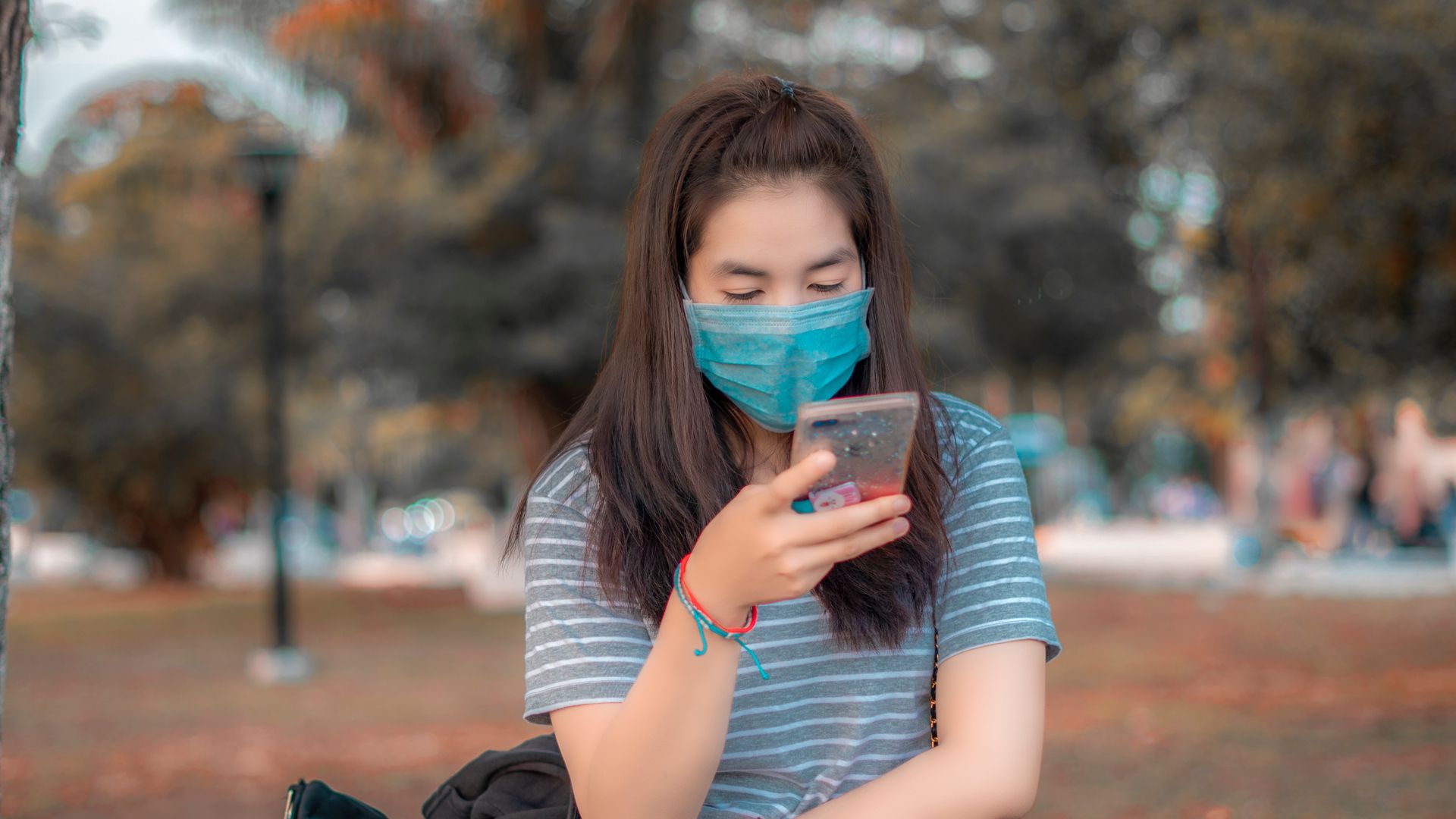 Woman wearing a face mask and texting on her phone in a park setting during summer.