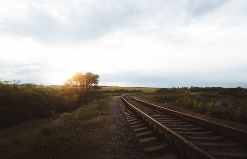 Free Empty narrow metal railway going along meadow with green lush bushes in nature with bright sunlight shining through tree against cloudy sky Stock Photo