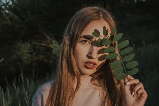 A woman poses with a leaf covering her eye, basking in sunlight outdoors amidst greenery.