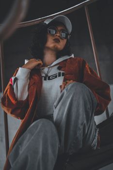 Young woman in street style posing with a moody look indoors wearing a cap and sunglasses.
