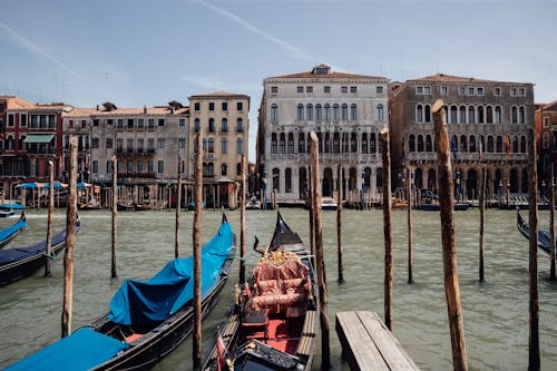 Gondolas moored on Grand Canal with wooden poles against facades of aged shabby residential buildings and blue sky in Venice