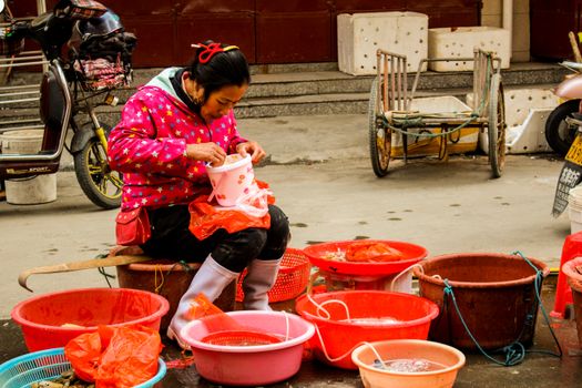Asian woman vendor preparing goods at an outdoor market, surrounded by colorful buckets.