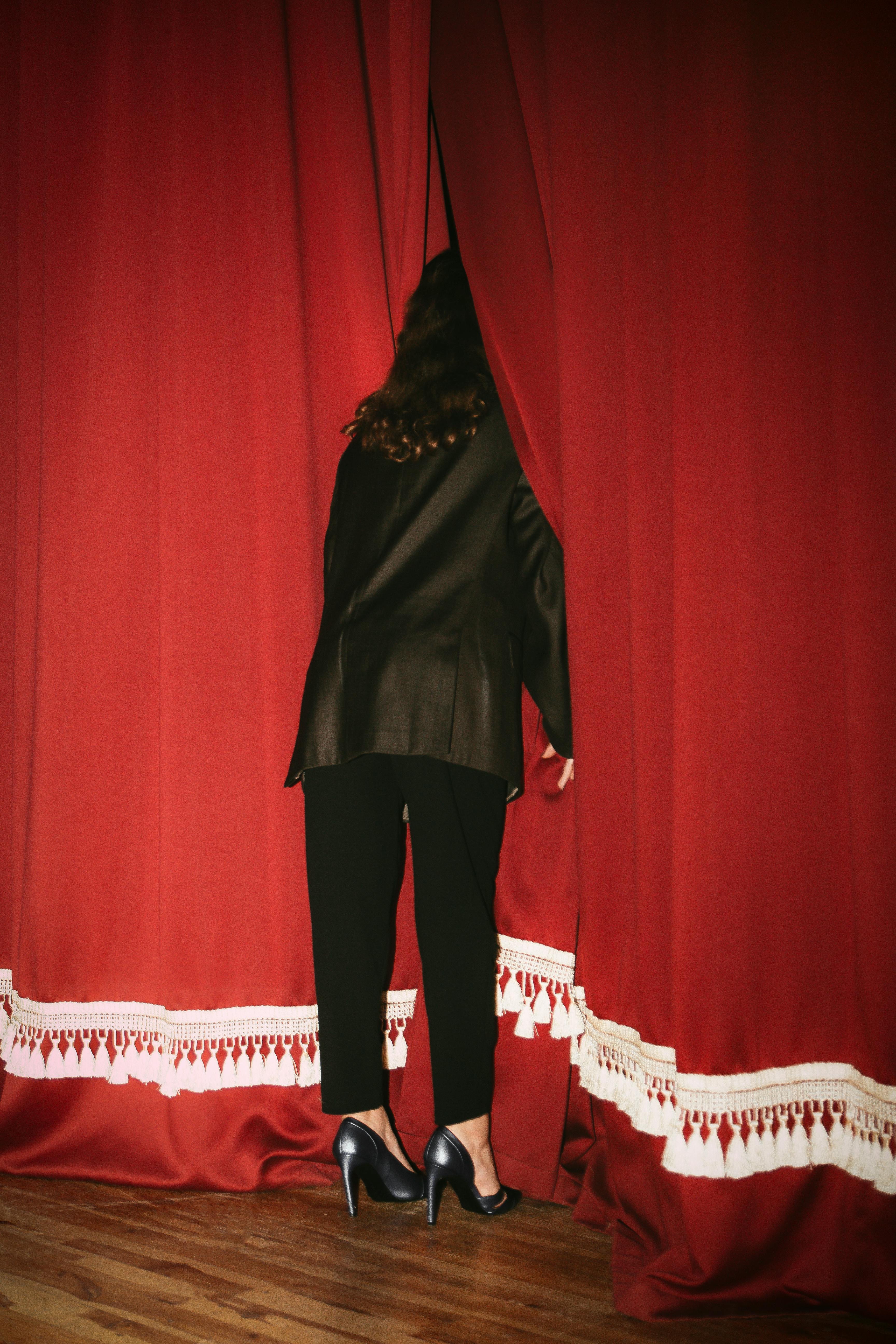 Free A woman peeks between red curtains, wearing black clothes in a theater setting. Stock Photo