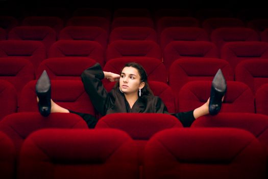 A woman in a black blazer and high heels relaxes on red theater seats in an empty auditorium.