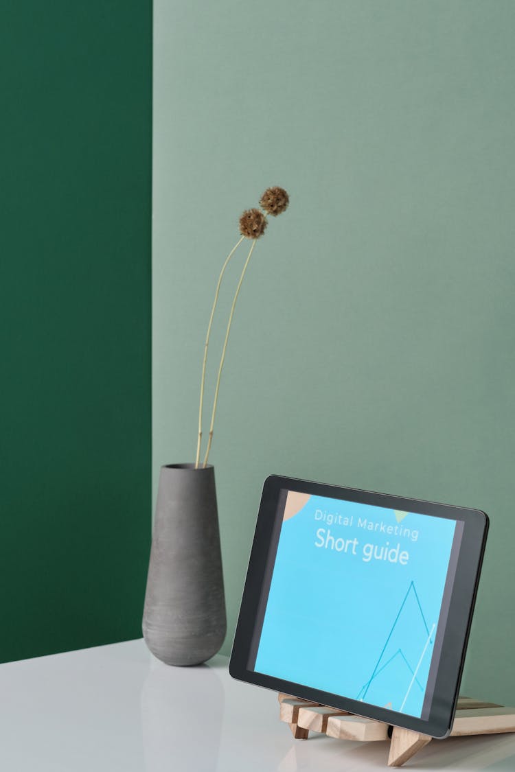 A Tablet With Short Guide Title On A Wooden Stand Beside Gray Vase With Flowers