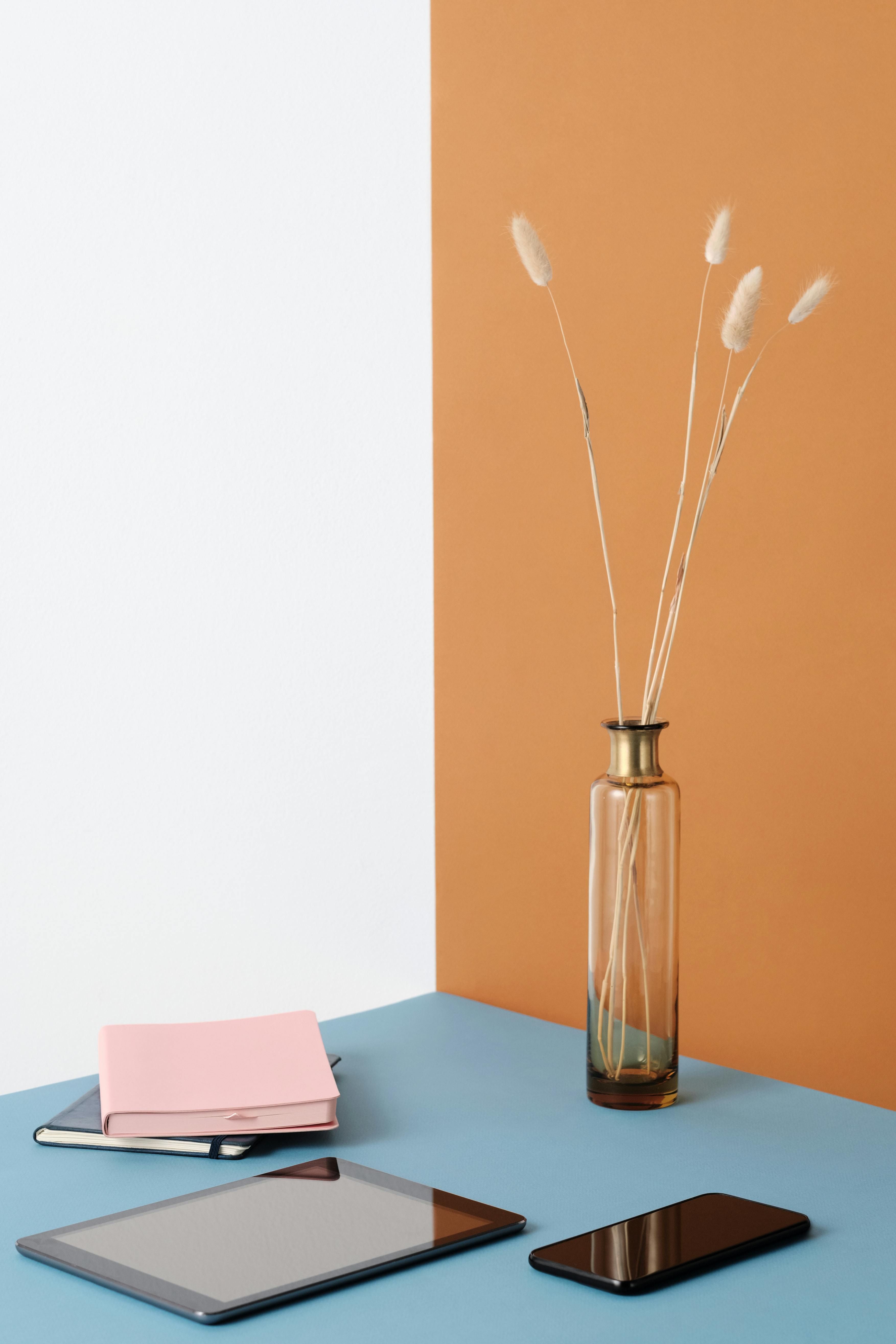 Free Stylish workspace setup featuring modern gadgets and a decorative glass vase with dried grass on a colorful surface. Stock Photo