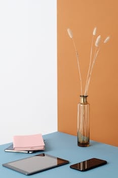 Stylish workspace setup featuring modern gadgets and a decorative glass vase with dried grass on a colorful surface.