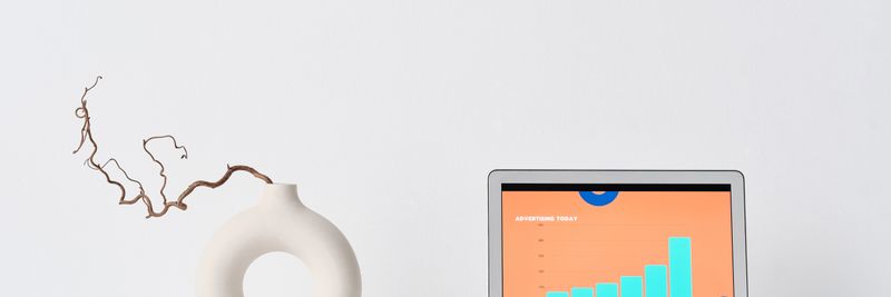 A minimalist workspace with a laptop, ceramic vase, and eyeglasses on a white desk.