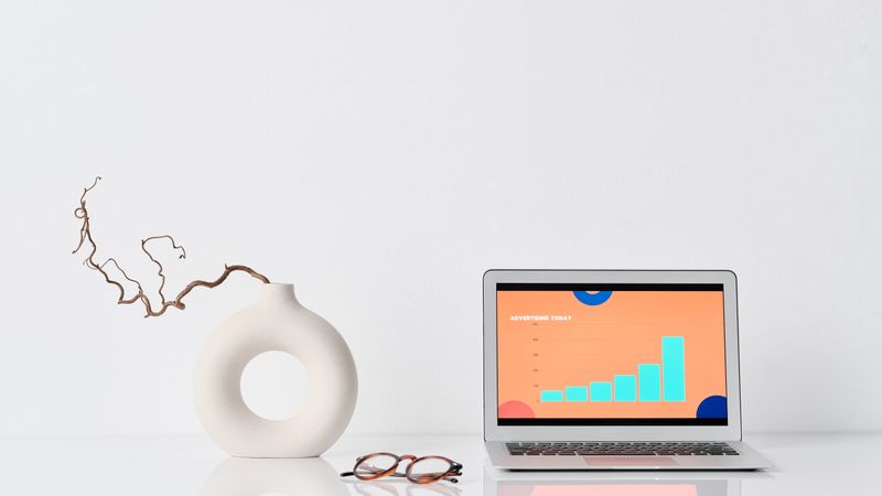 A minimalist workspace with a laptop, ceramic vase, and eyeglasses on a white desk.