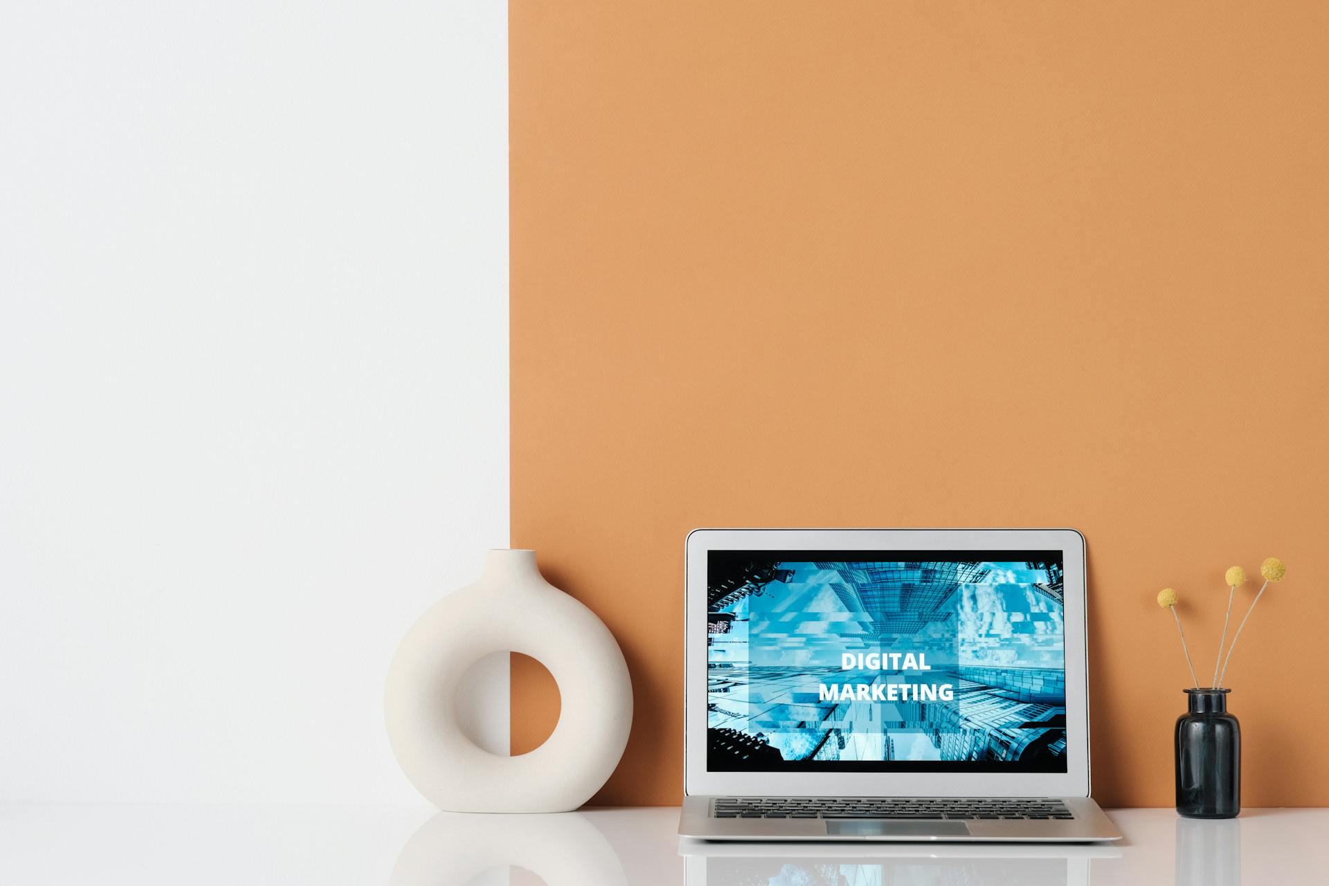 Minimalist workspace featuring a laptop with digital marketing text, a ceramic vase, and dried grass.