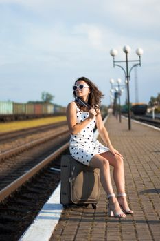 Stylish woman in white polka dot dress sitting on luggage at railway station platform.