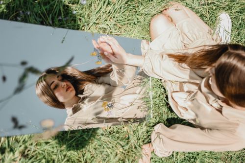 Free A woman in a beige outfit sits in grass, reflected in a mirror outdoors. Stock Photo