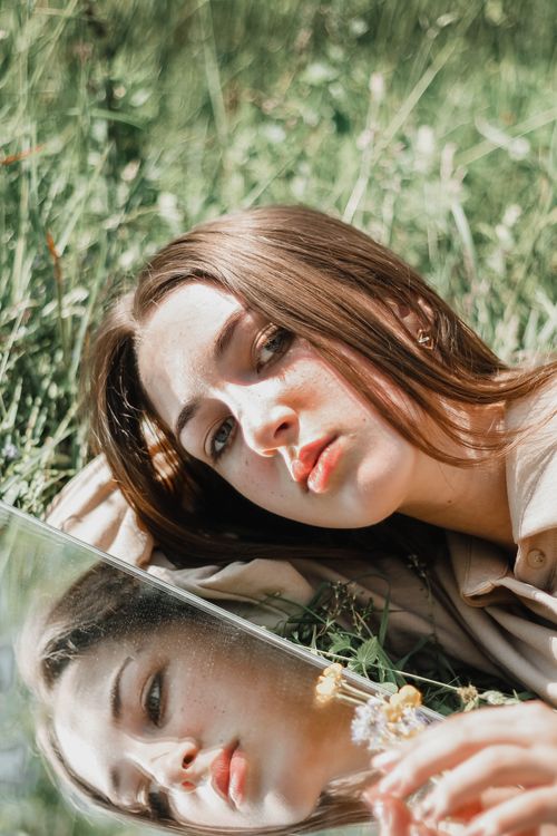 Free Artistic portrait of a woman lying with a mirror in a grassy field reflecting her face. Stock Photo