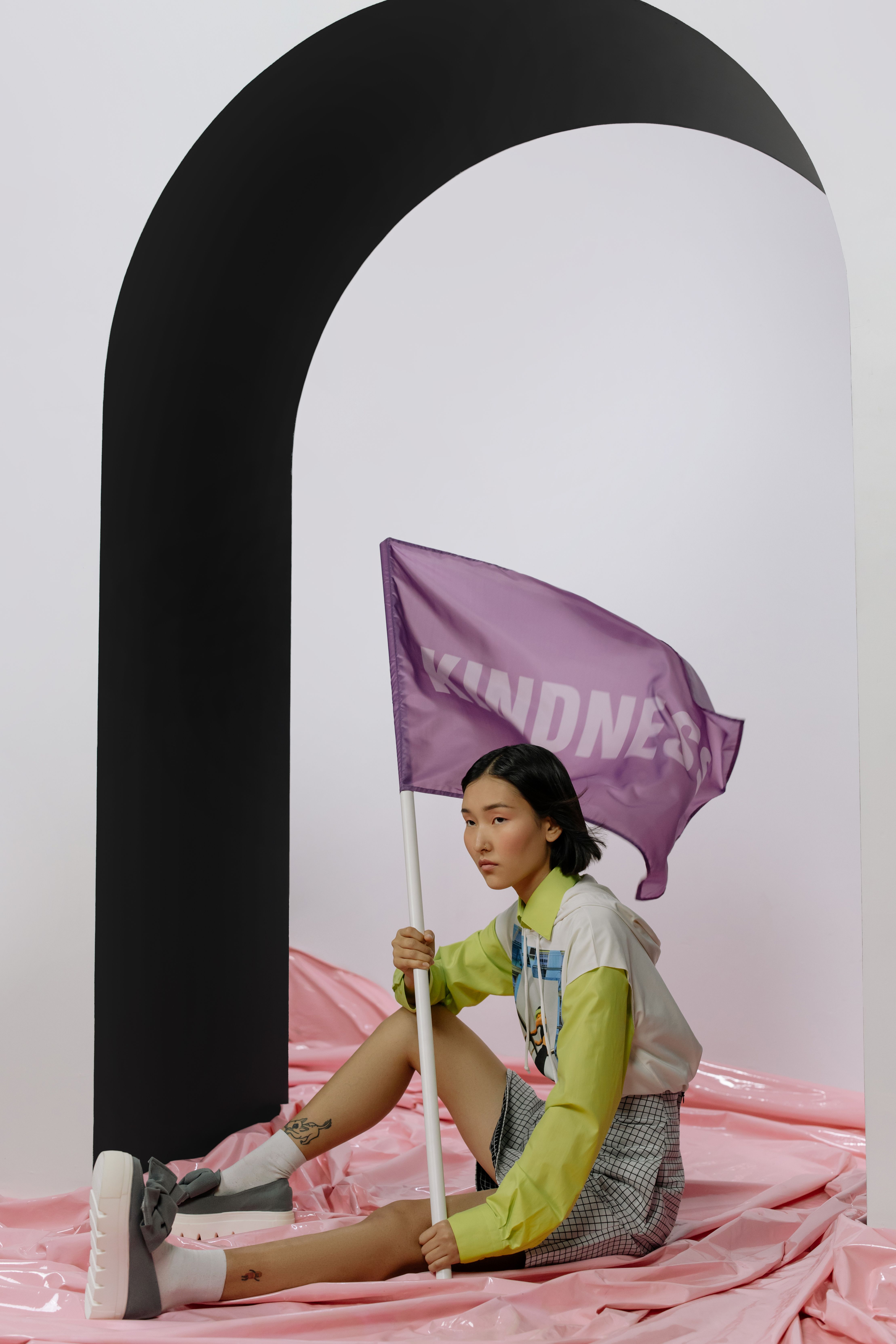 Asian woman holding a purple flag with 'Kindness', set against artistic arch in studio.