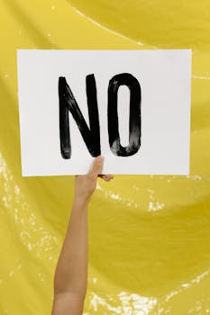 A hand holding a 'NO' sign against a bright yellow background.