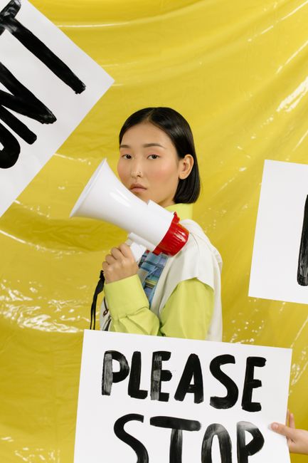 Asian woman holding a megaphone with protest signs in a vibrant yellow background